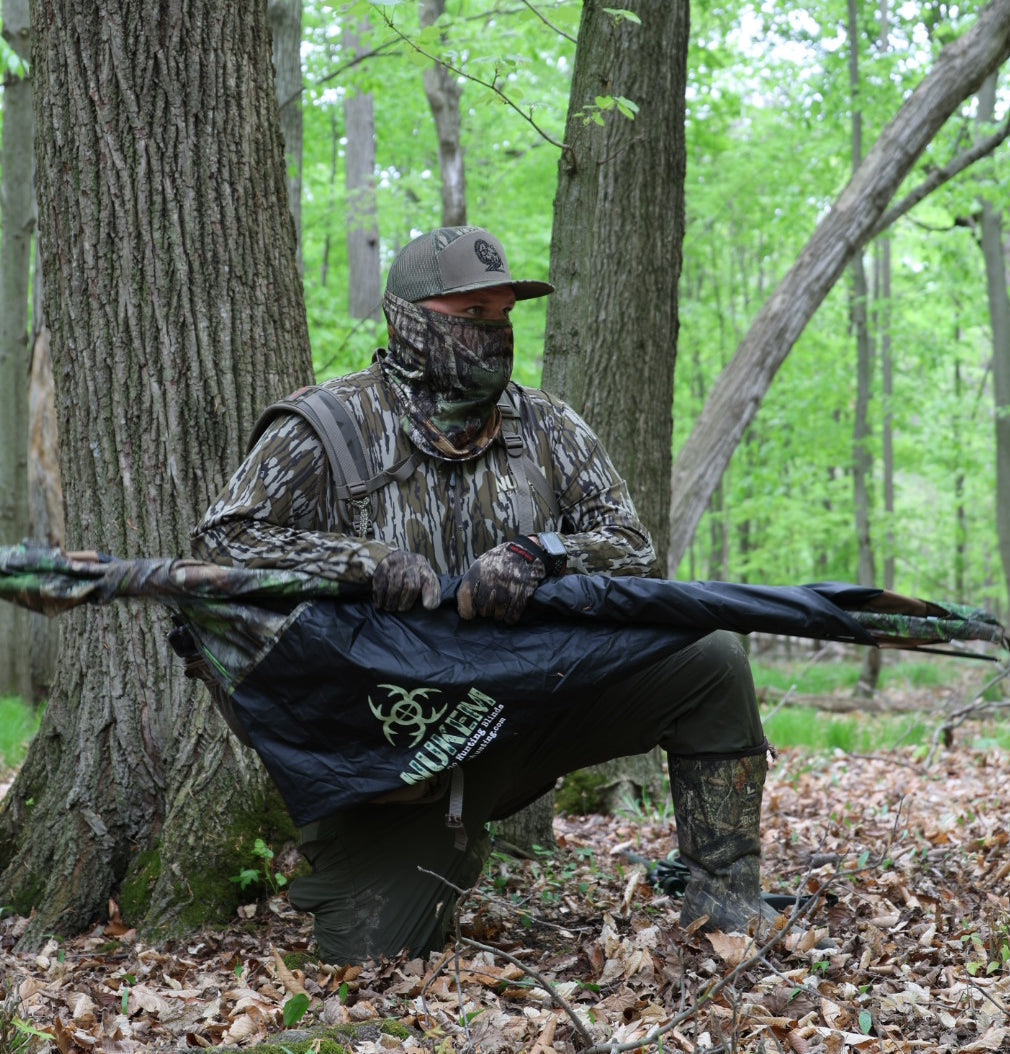 Hunter in woodland camo gear holding bow in forest, wearing face mask and gloves for concealment