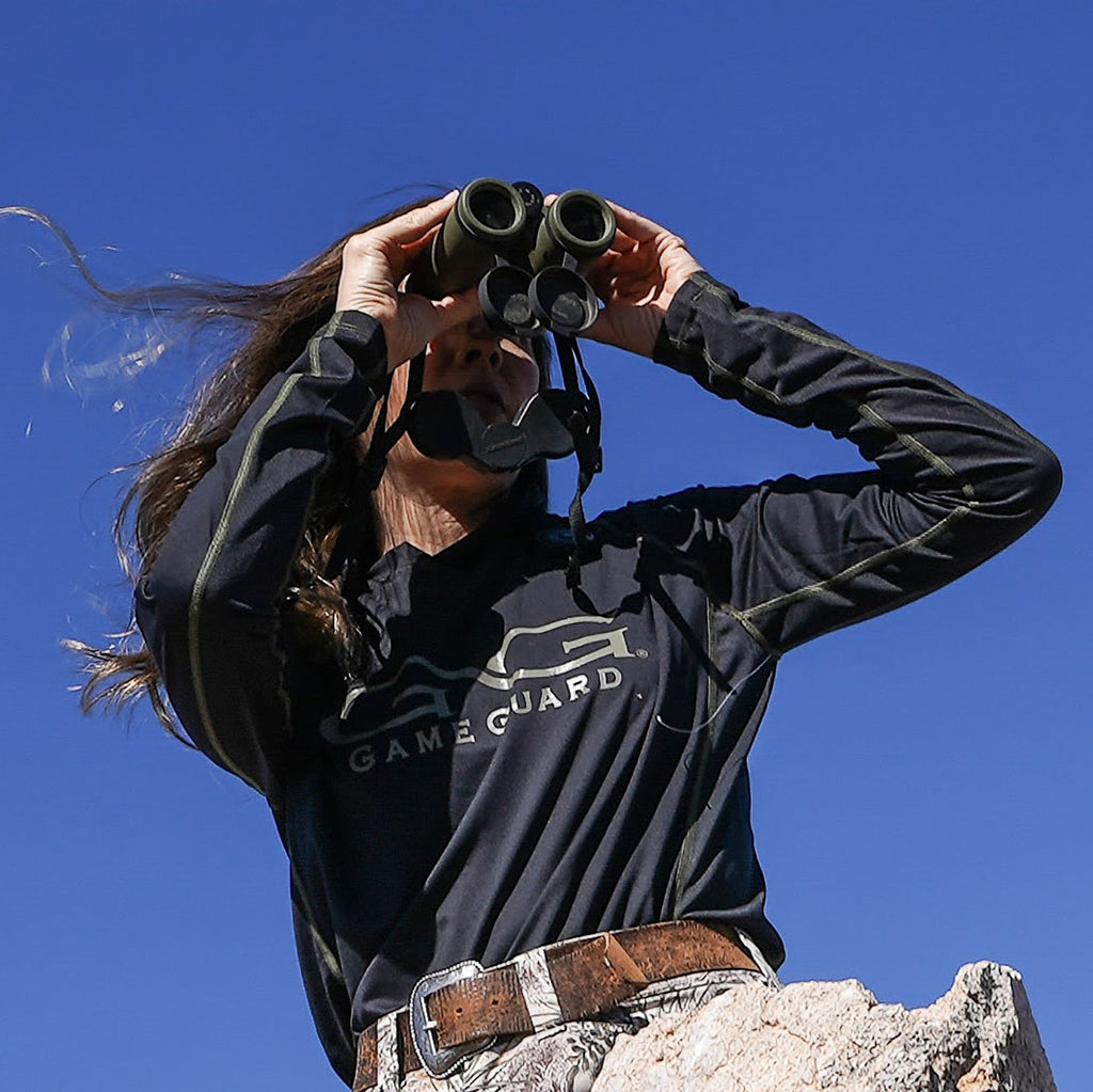 Woman using binoculars outdoors in Game Guard hunting apparel under clear blue sky