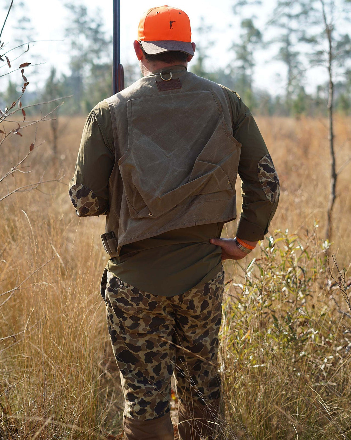 Hunter in a blaze orange cap and Original Camo Field Pants walking into a pine field