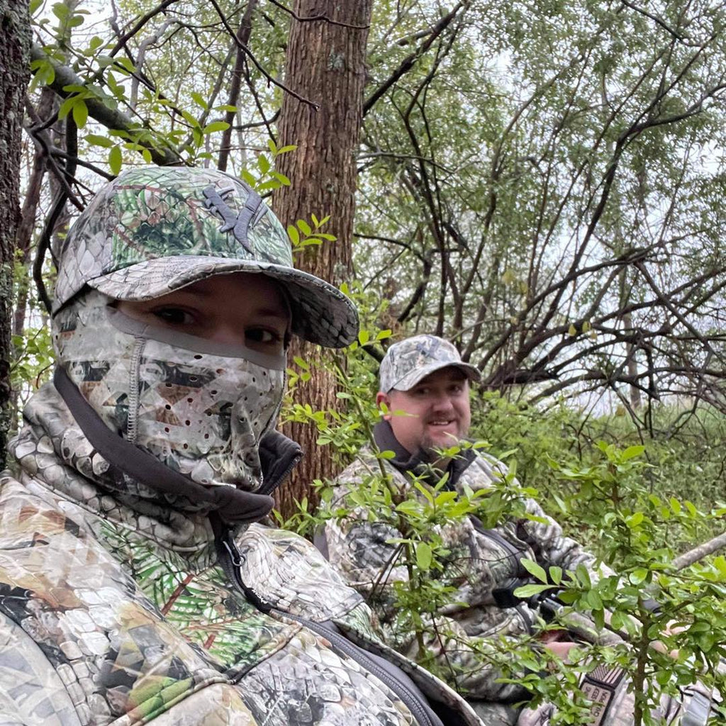 Two hunters in a ground blind, one wearing a Vycah camo face mask and cap