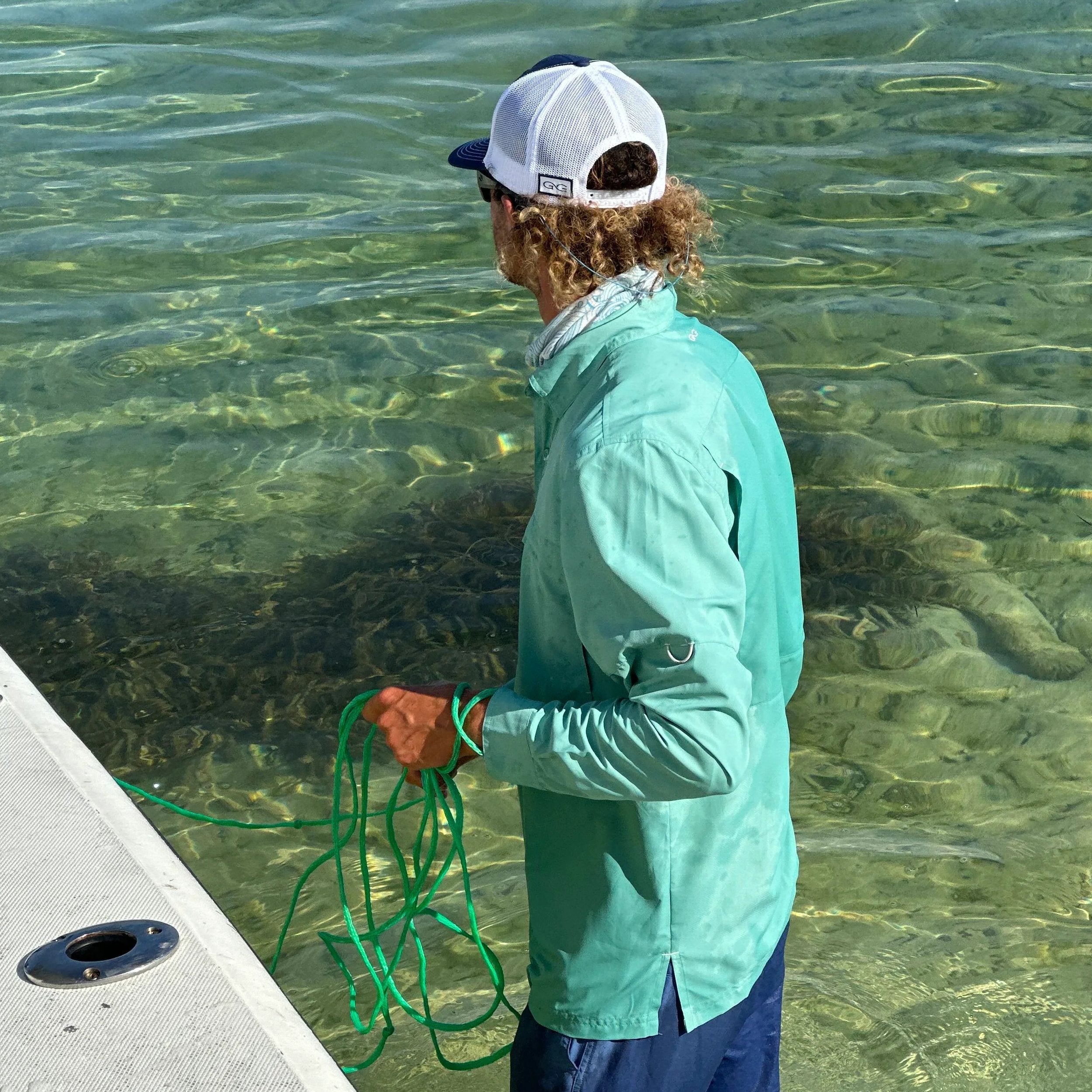 Man in teal fishing shirt and cap holding green rope on boat in clear water