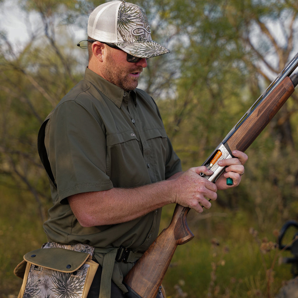 Hunter in camo hat and olive shirt loading shotgun in outdoor setting
