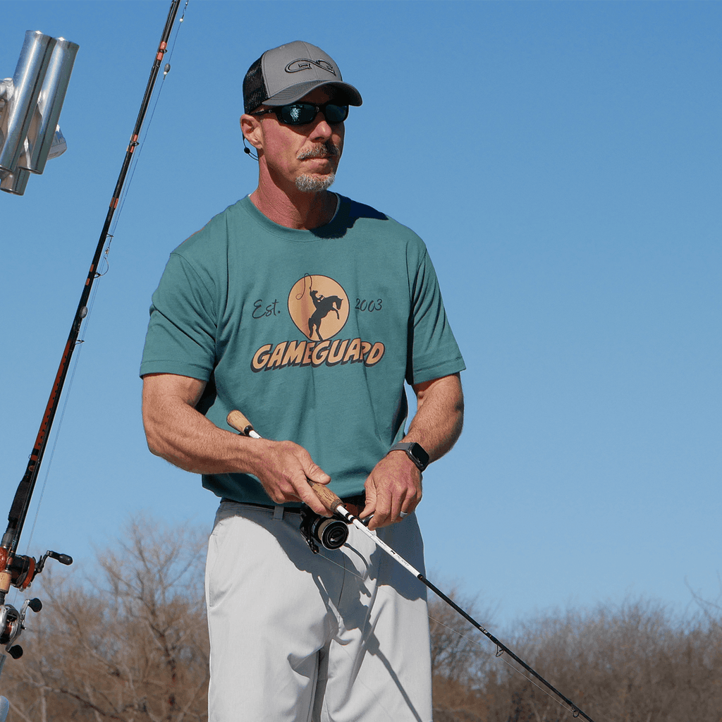 Man fishing in teal GameGuard t-shirt, gray cap, and sunglasses, holding rod under blue sky