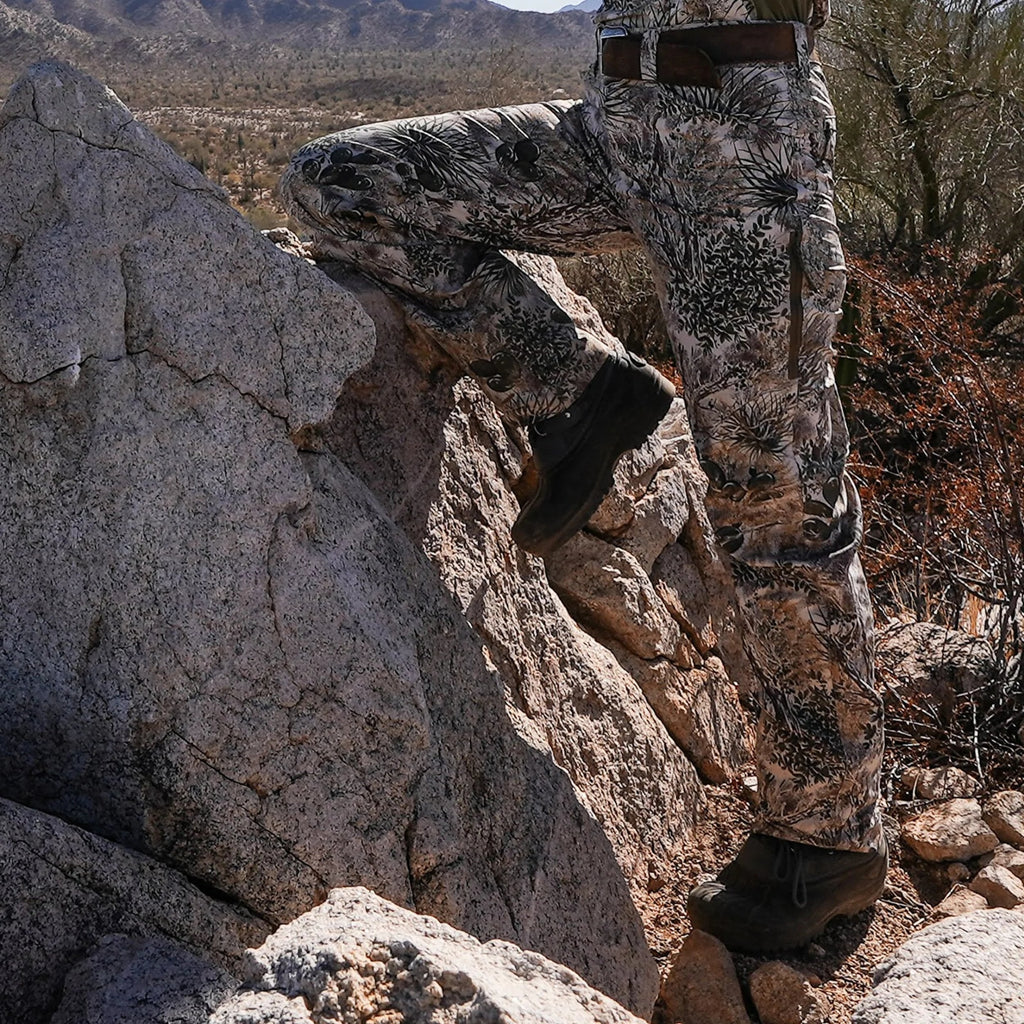 Desert camo hunting pants with cactus print, worn on rocky terrain for outdoor stealth