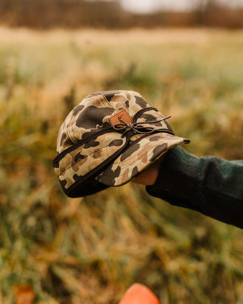 Original Camo Stormy Kromer cap held in a hand in tall grass