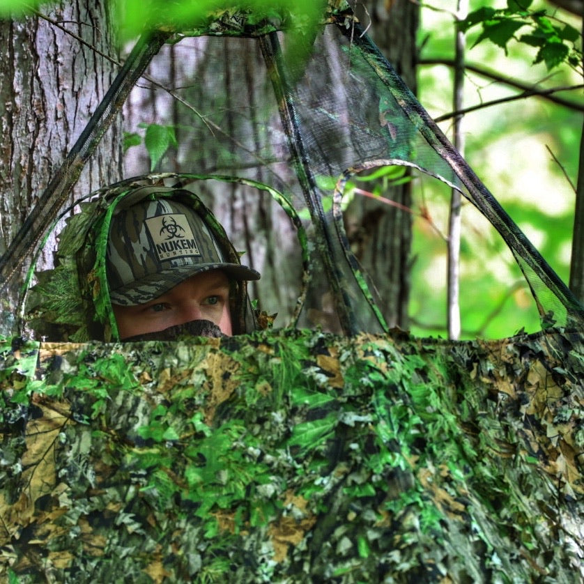 Hunter in leafy camouflage blind with Nukem hat, blending into forest for deer hunting