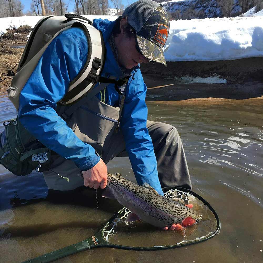 Angler in blue waterproof jacket holding rainbow trout in net, wading in snowy river, camo hat