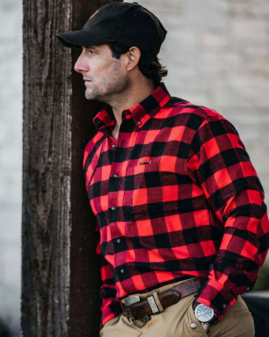 Man in a black cap wearing the red Buffalo Plaid Sportsmans Shirt leaning on a post
