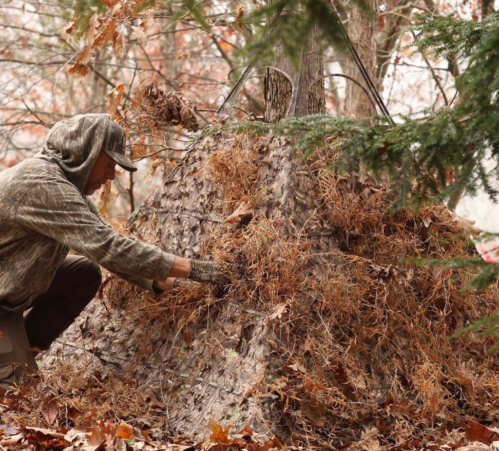 Hunter setting up camo ground blind with leafy concealment in autumn woods for deer hunting