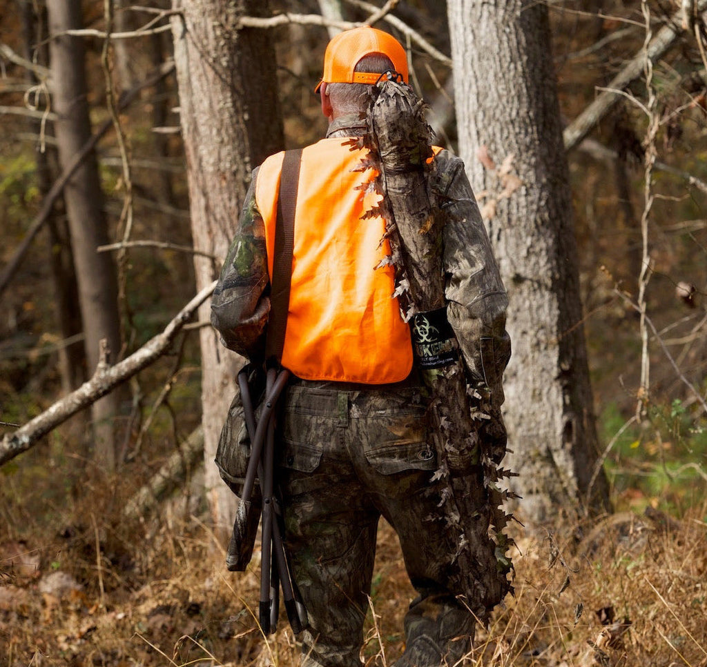 Hunter in camo gear with orange safety vest and leafy ghillie suit walking through woods