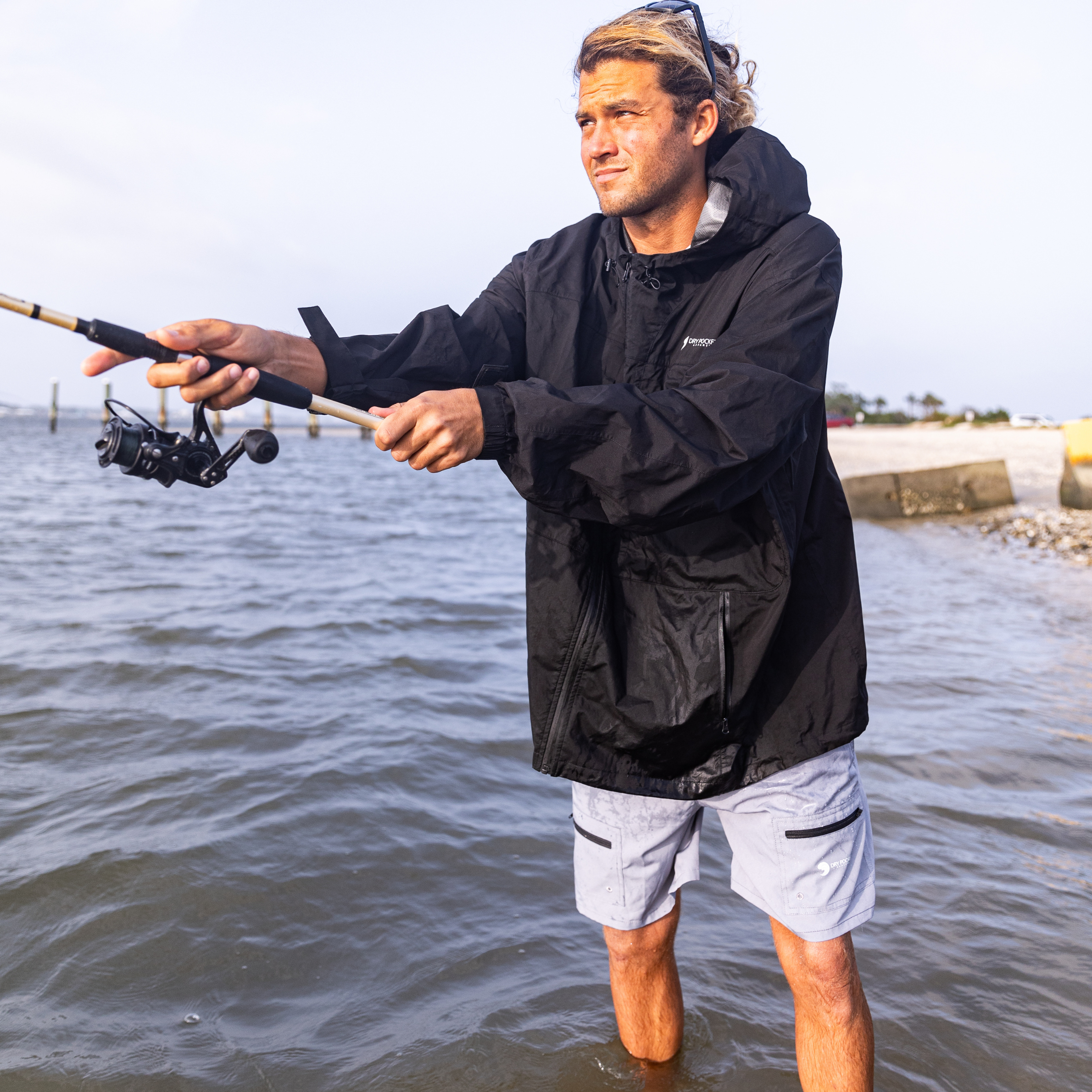 Man fishing in black waterproof jacket and gray shorts at shoreline, casting rod in water
