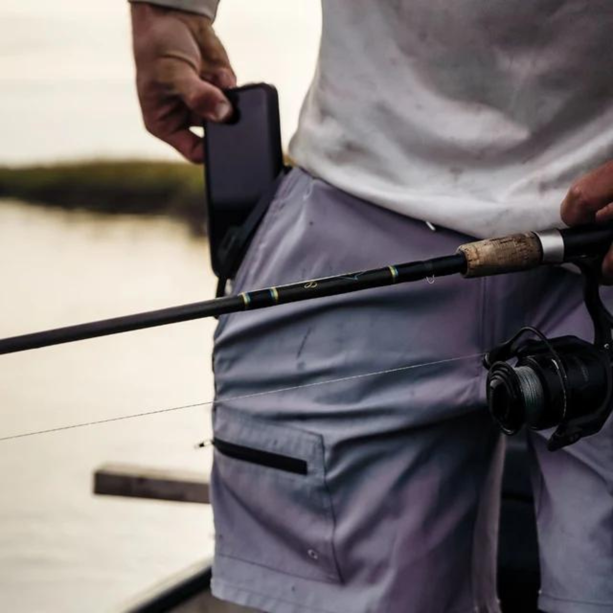 Angler holding fishing rod with reel, wearing gray shorts, on boat near water