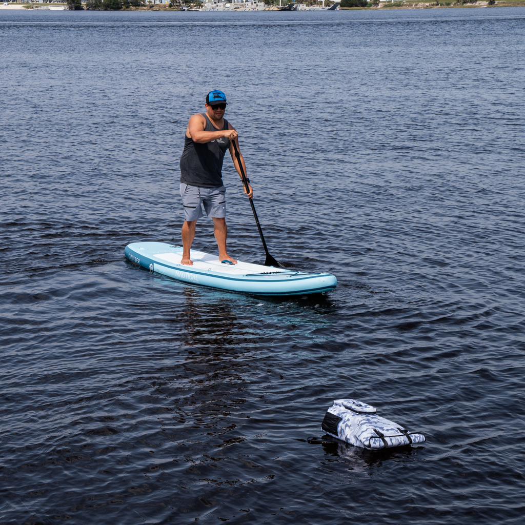 Man paddleboarding on light blue inflatable SUP with camo dry bag, ideal for fishing and outdoor adventures.