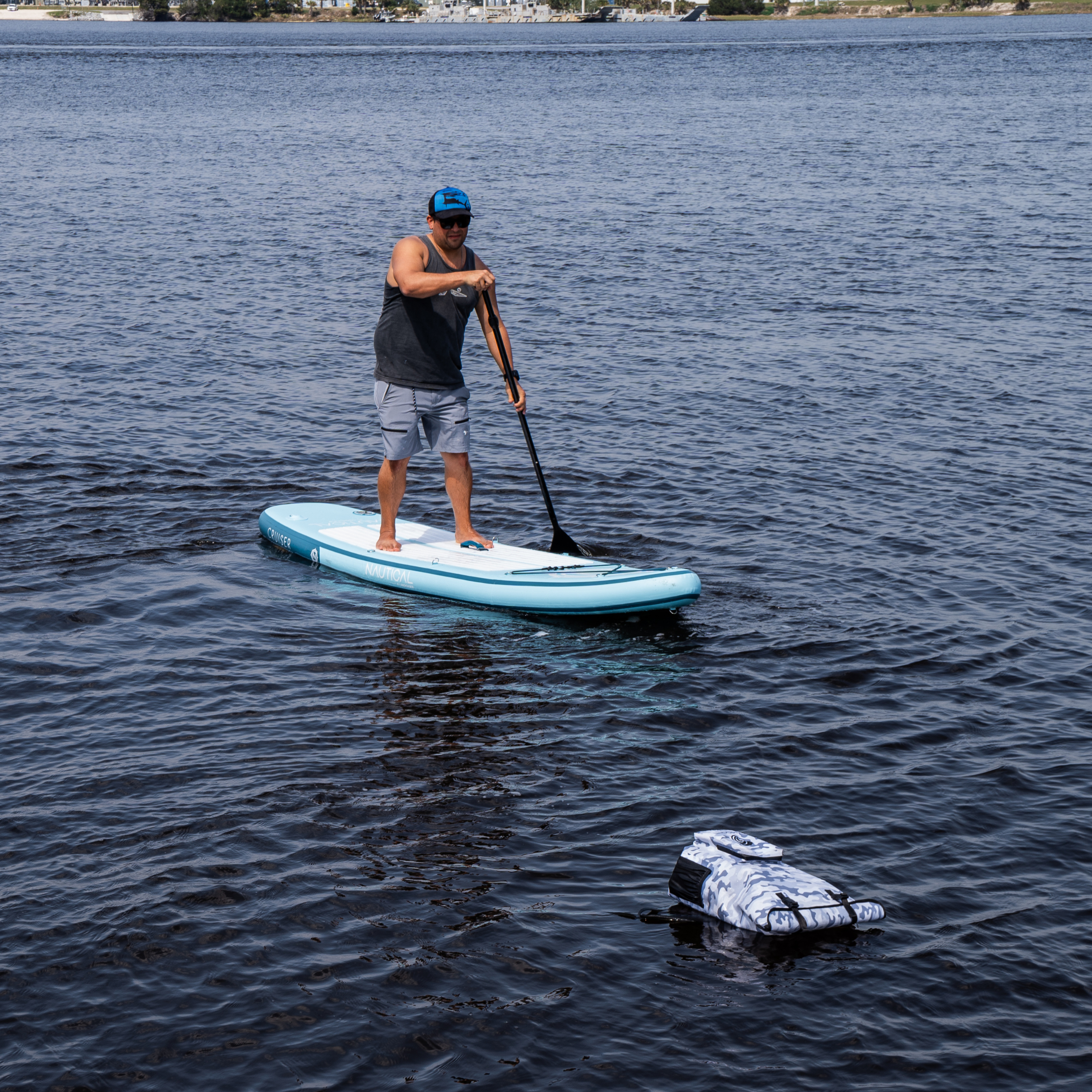 Man paddleboarding on light blue inflatable SUP with camo dry bag, ideal for fishing and outdoor adventures.