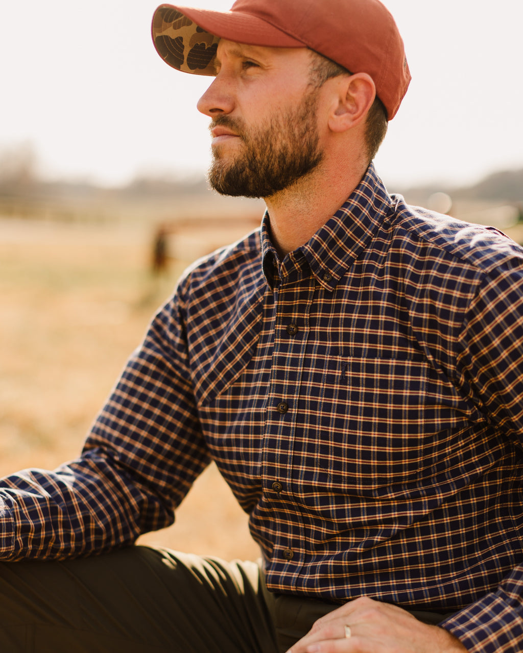 Man in the Burnt Orange Arthur Core Hat and a check shirt sitting by a hay bale