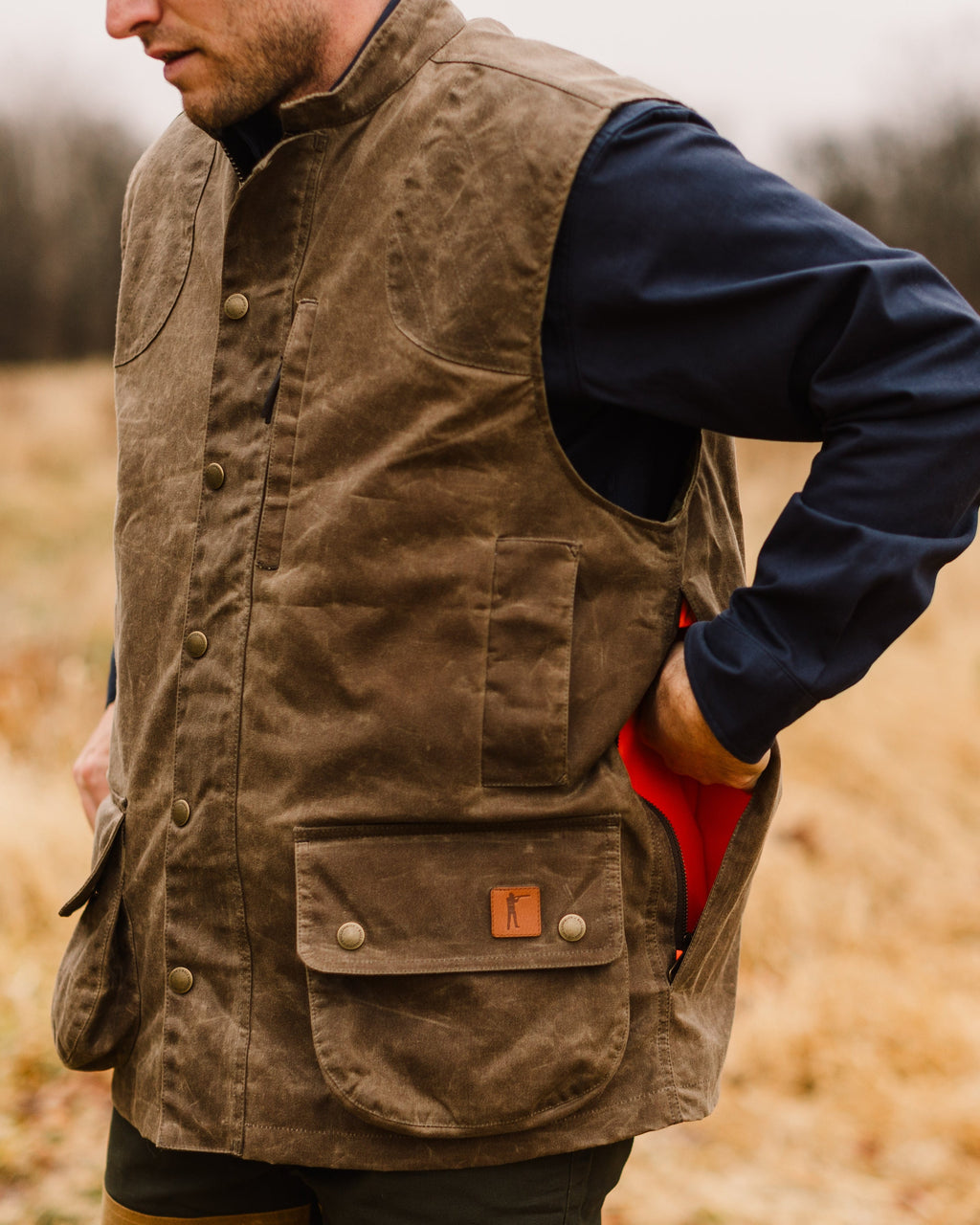 Dark Oak Upland Vest worn over a navy shirt, hand in the blaze orange lined game pocket