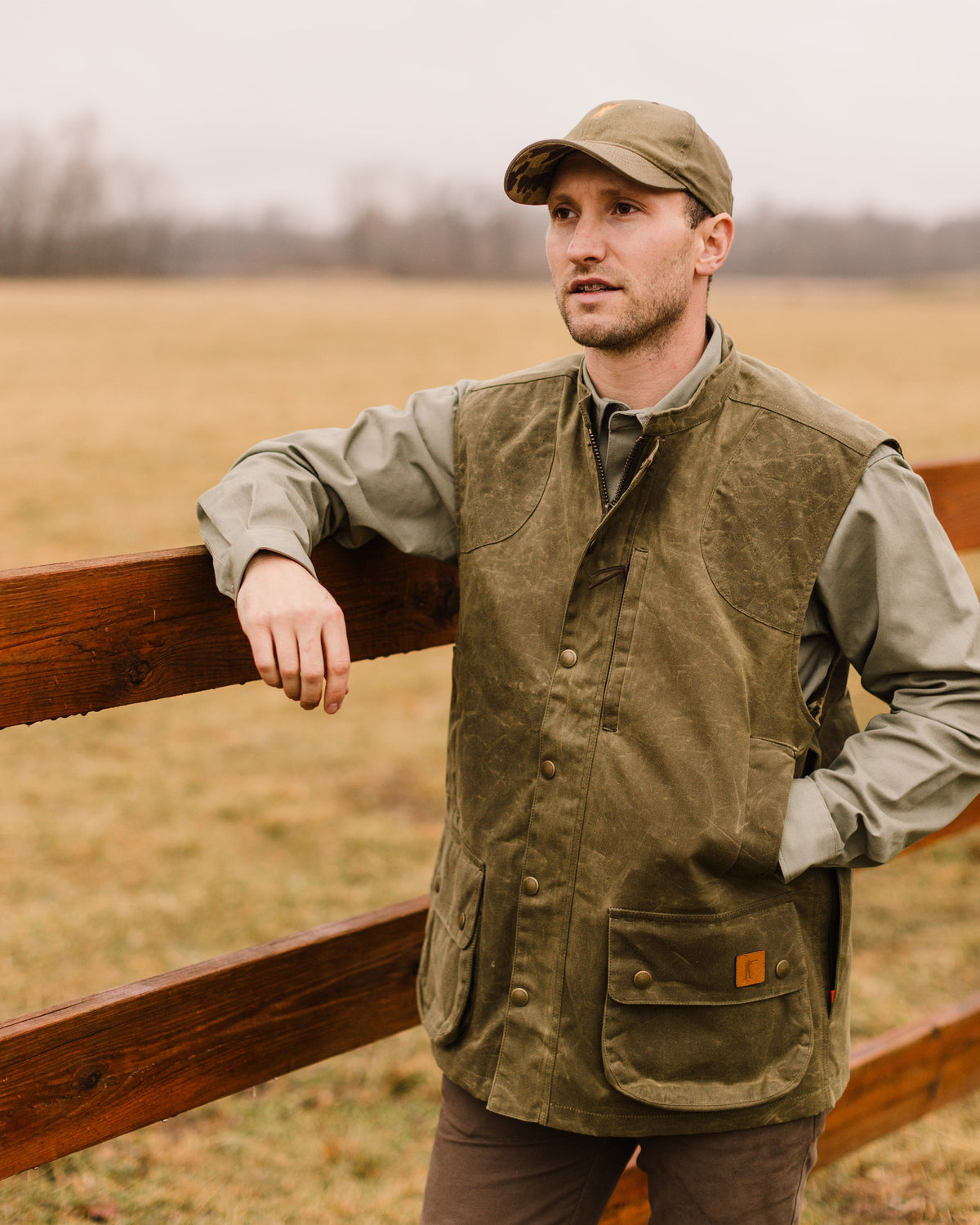 Man leaning on a split rail fence wearing the Olive Upland Vest and a field cap