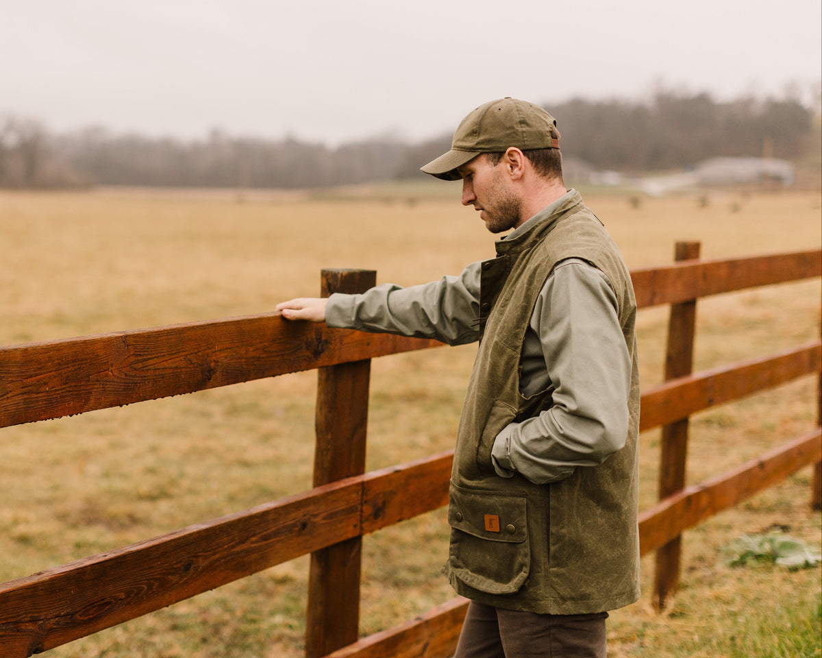 Man in the Olive Upland Vest standing at a wooden fence line in an open field