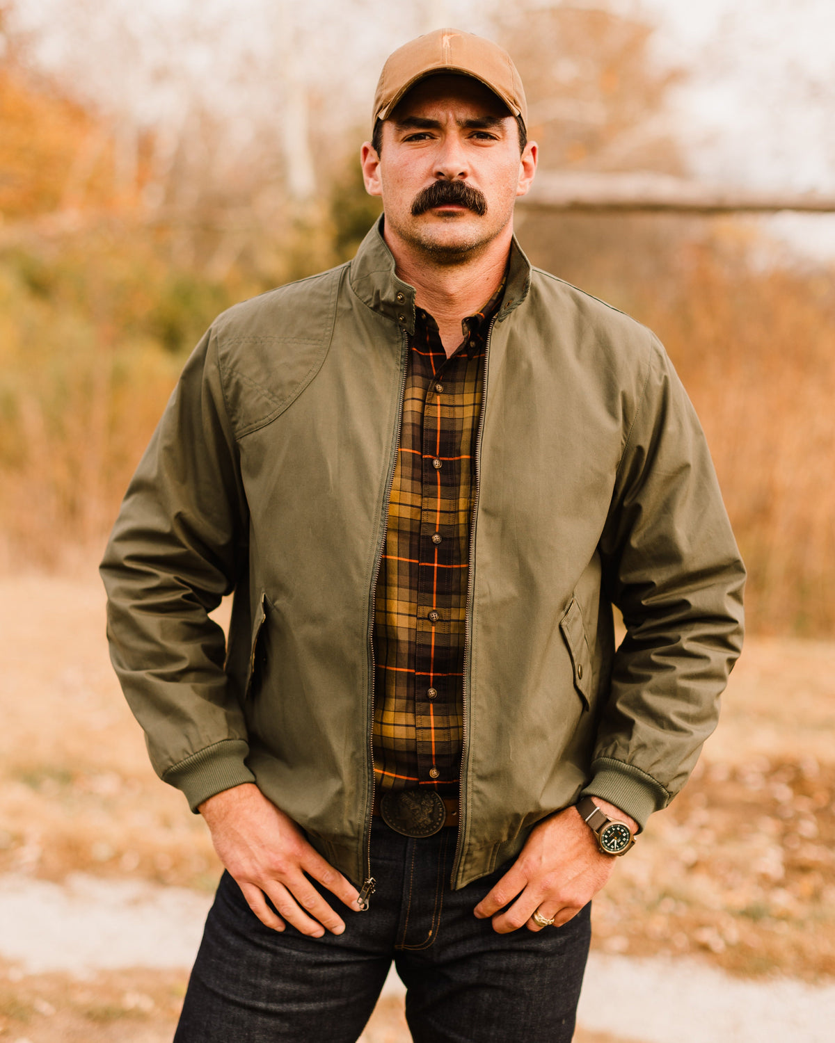 Man in a cap wearing the Sage Green Harrington Jacket over a plaid flannel in a fall field