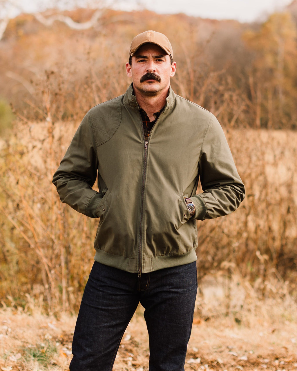 Man standing in tall grass wearing the Sage Green Harrington Jacket