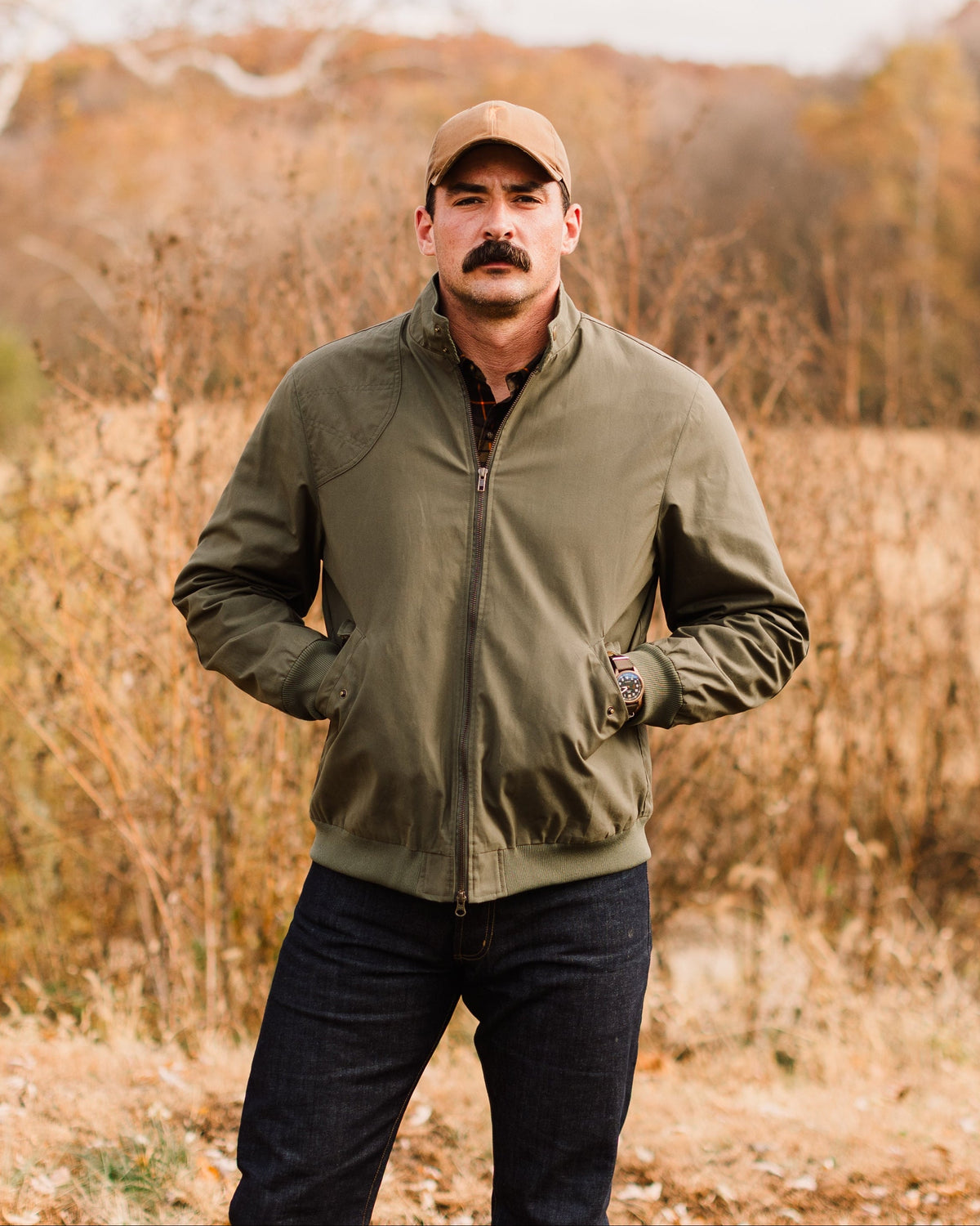Man standing in tall grass wearing the Sage Green Harrington Jacket