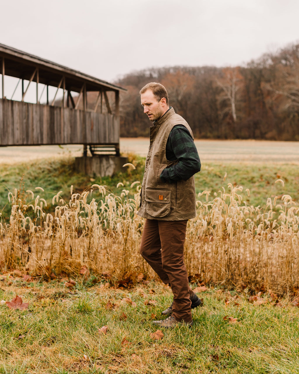 Hunter wearing the Ball and Buck Upland Vest in Dark Oak walking through tall grass near an old barn