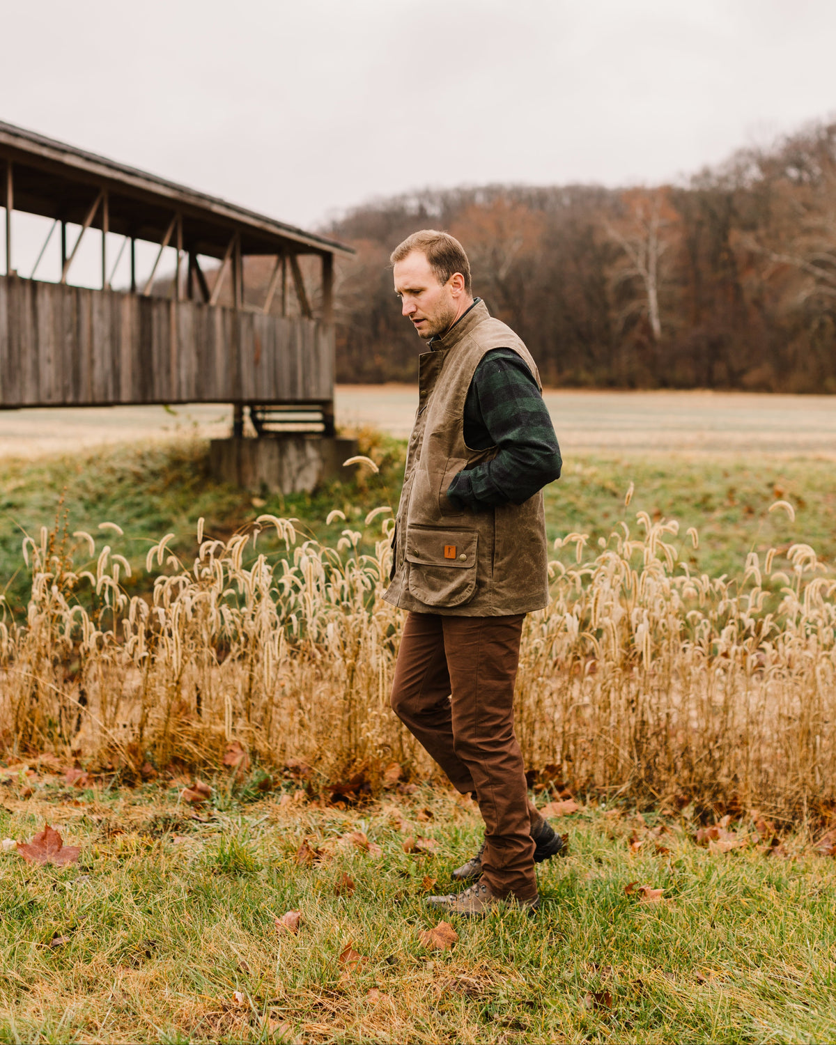 Hunter wearing the Ball and Buck Upland Vest in Dark Oak walking through tall grass near an old barn