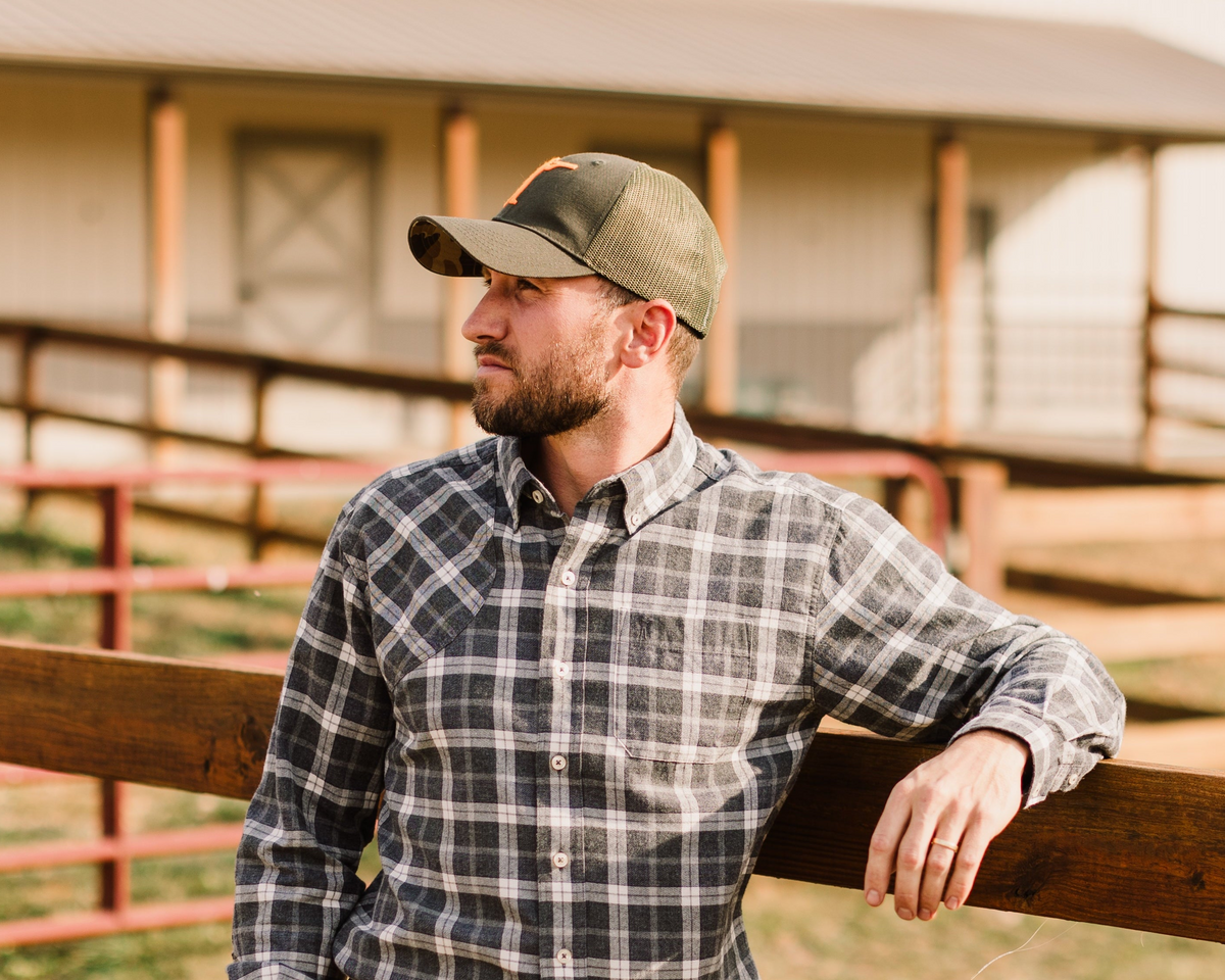 Man leaning on a fence in a plaid shirt and Arthur Core Snapback