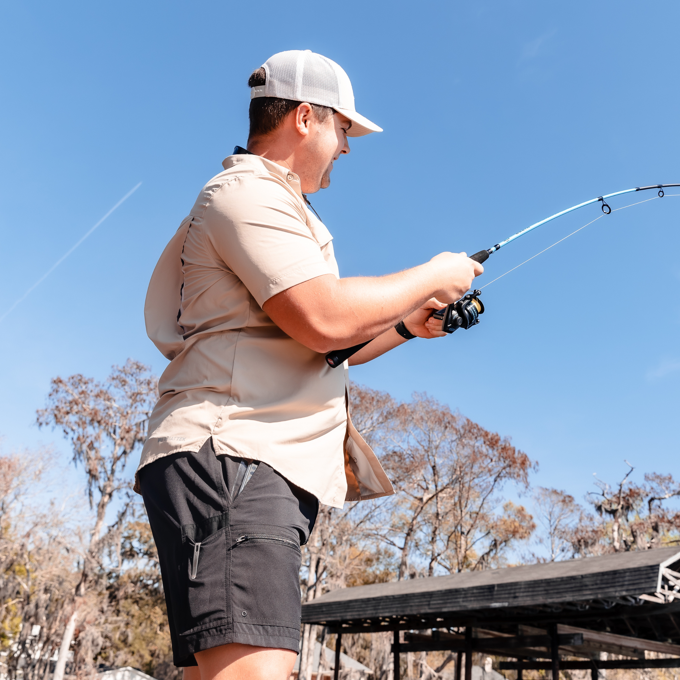 Man fishing with light blue rod, wearing tan shirt and black shorts under clear sky