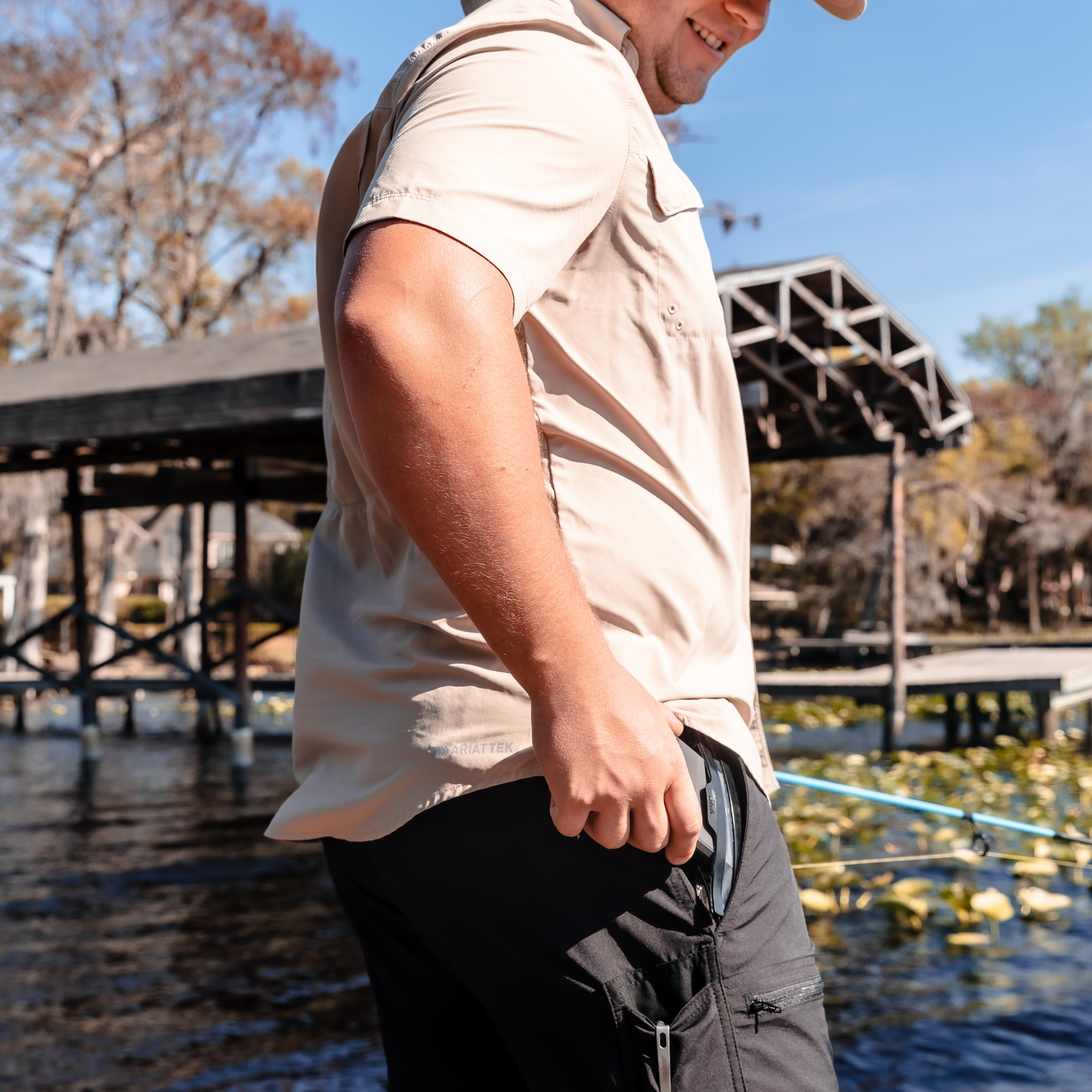 Tan short sleeve fishing shirt with Ariattek logo, worn by man on dock near water, ideal for outdoor adventures.