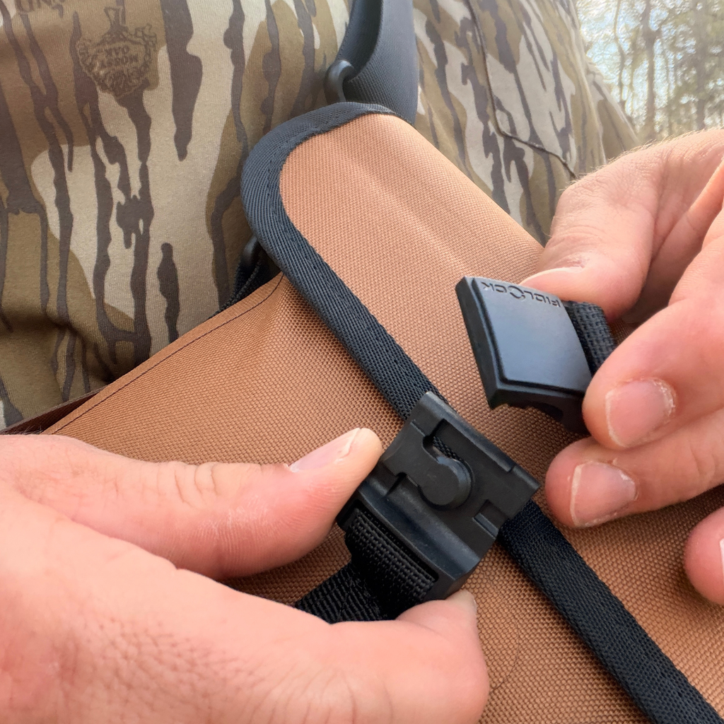 Hands adjusting black Fidlock magnetic buckle on brown hunting gear strap, worn over camo apparel.