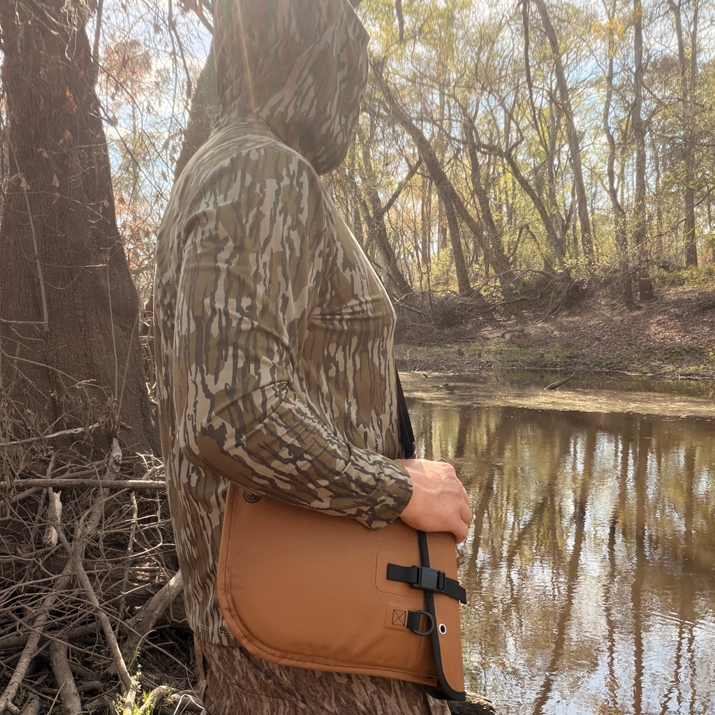 Camouflage hooded hunting jacket with tan utility pouch, worn by hunter near wooded pond