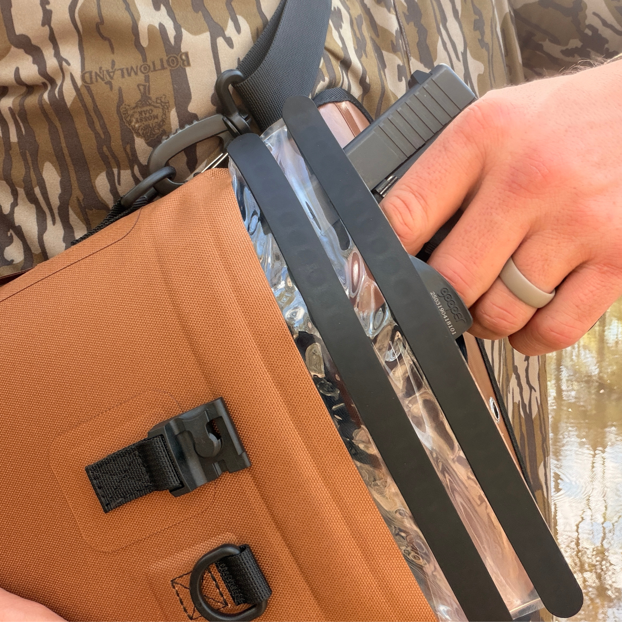 Hand holding clear tactical holster with black straps, worn over Mossy Oak camo gear, ideal for hunting or outdoor use