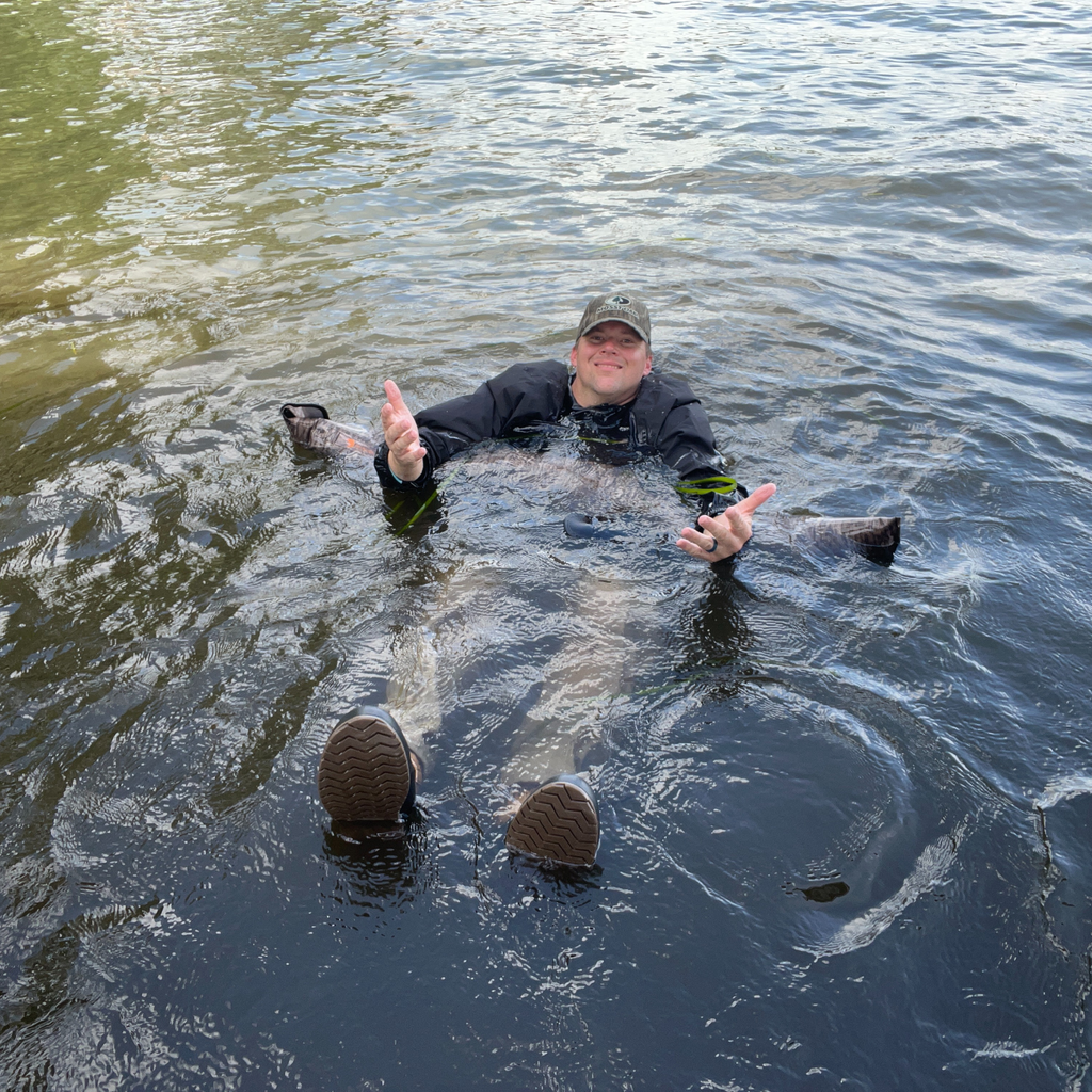 Man floating in river wearing waterproof waders and camo hat, testing hunting gear in water
