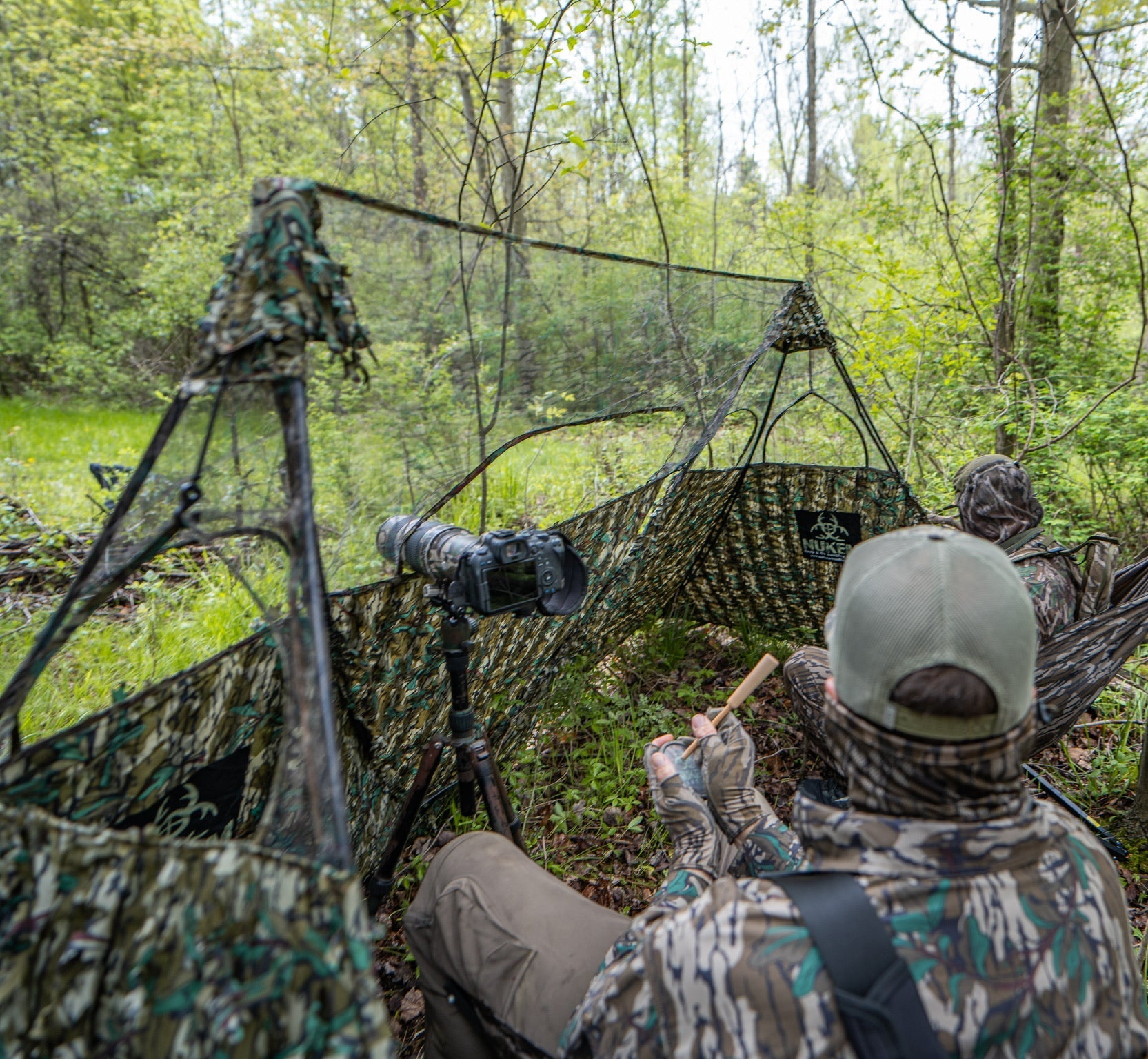 Camo hunting blind with photographer in forest, using camera on tripod for wildlife observation