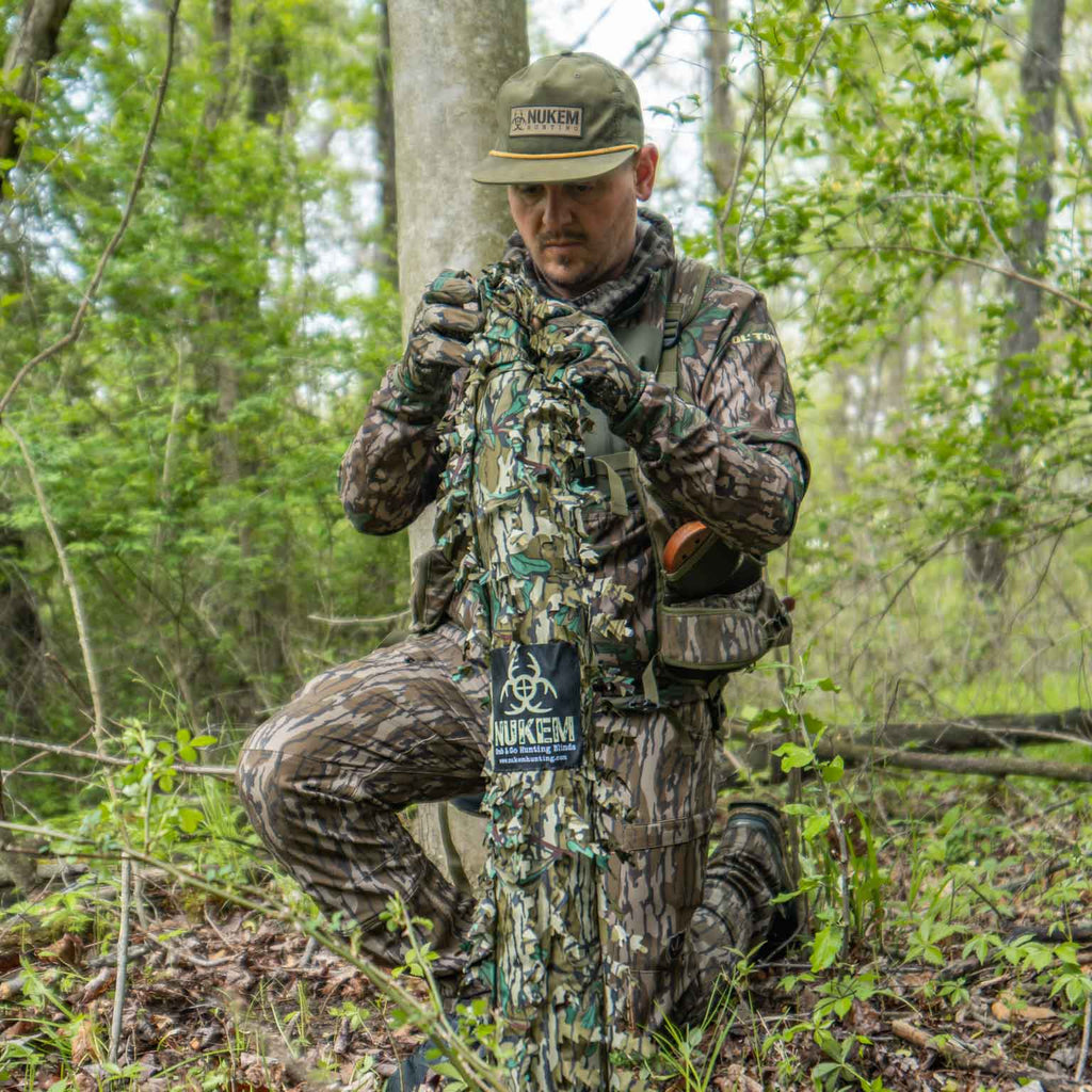 Hunter in Nukem camo gear adjusting ghillie suit in forest, ideal for deer hunting and concealment