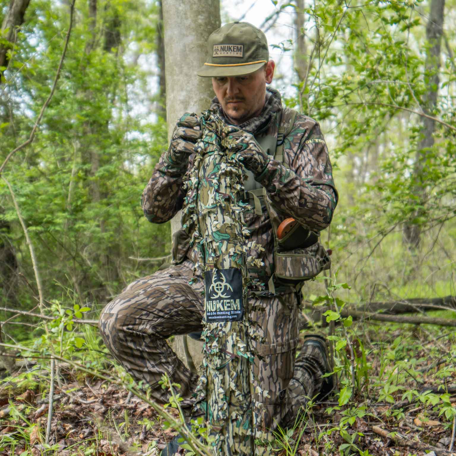 Hunter in Nukem camo gear adjusting ghillie suit in forest, ideal for deer hunting and concealment