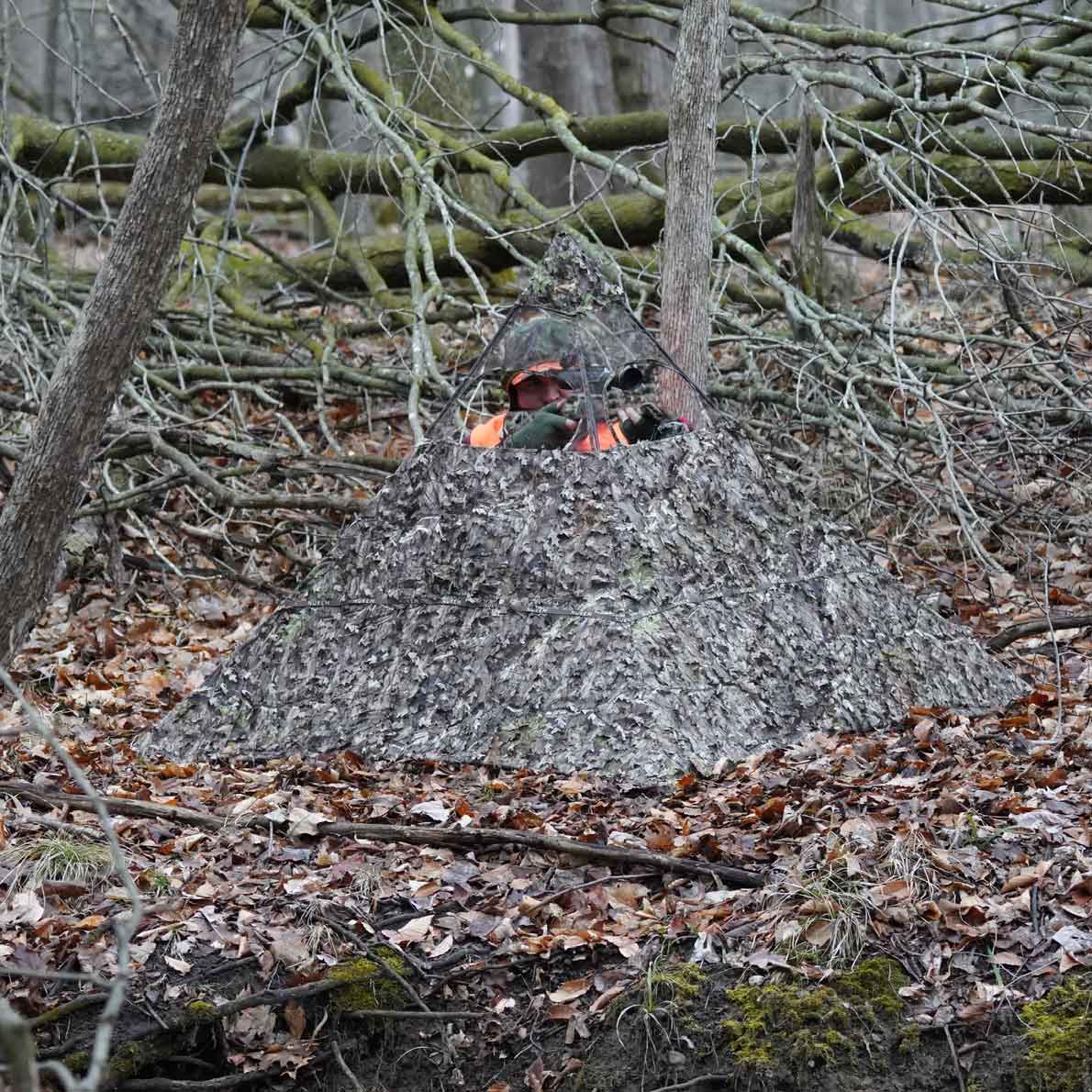 Camouflage hunting blind in forest, blending with fallen leaves and branches for concealment during deer hunting.