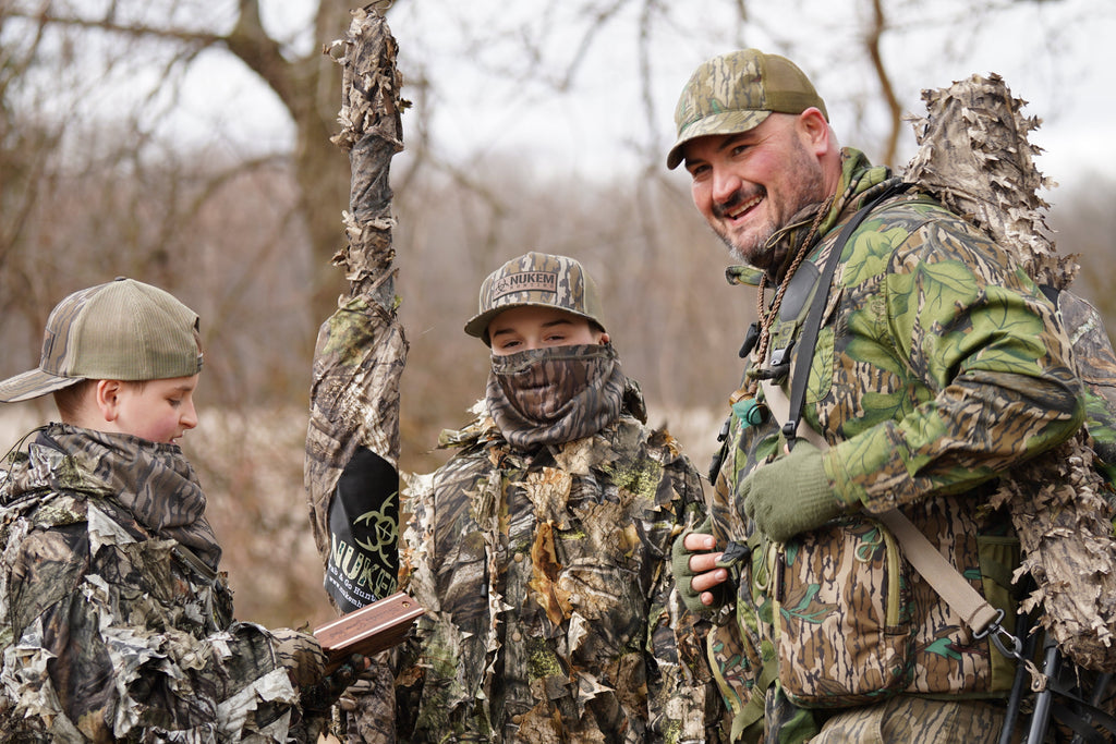 Hunters in camo gear with leafy ghillie suits and caps, outdoors in wooded setting
