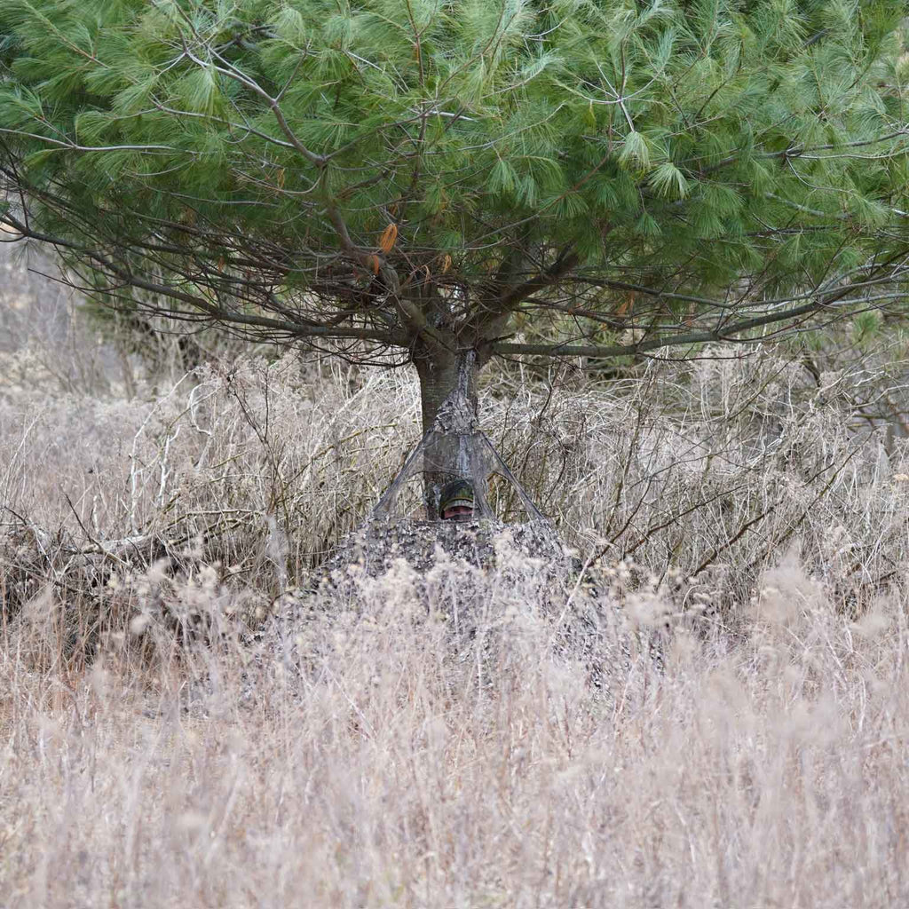 Hunter in camo ghillie suit blending into brush and pine tree for concealment during hunting