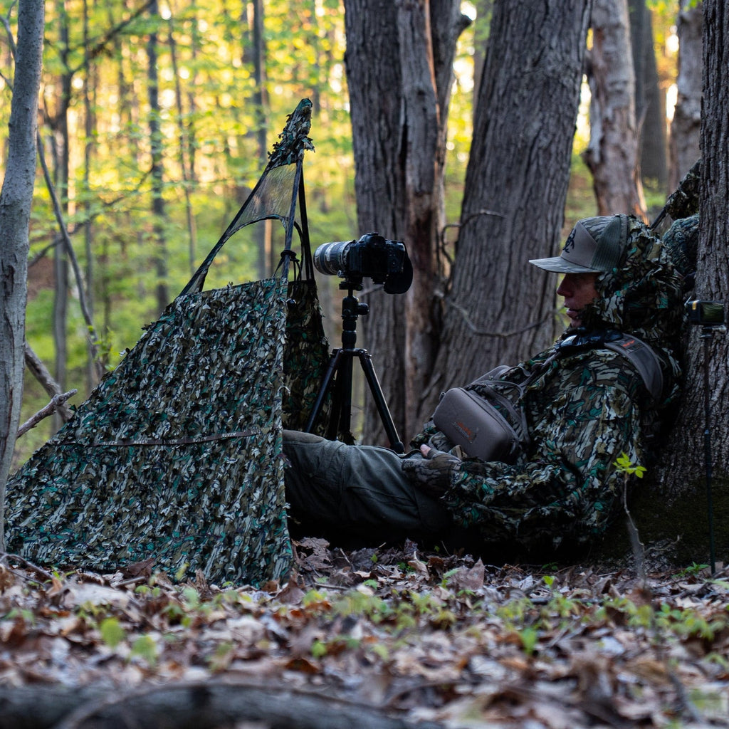 Hunter in leafy camo blind with camera setup in forest, ideal for wildlife photography and hunting concealment