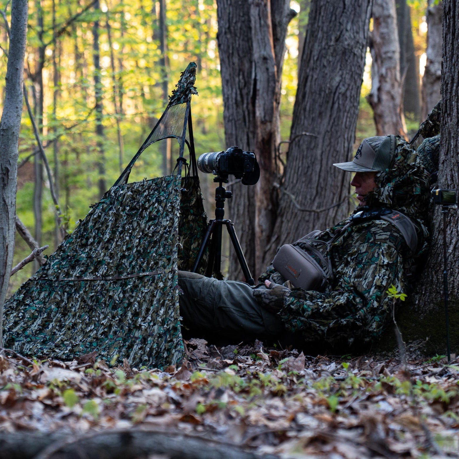 Hunter in leafy camo blind with camera setup in forest, ideal for wildlife photography and hunting concealment