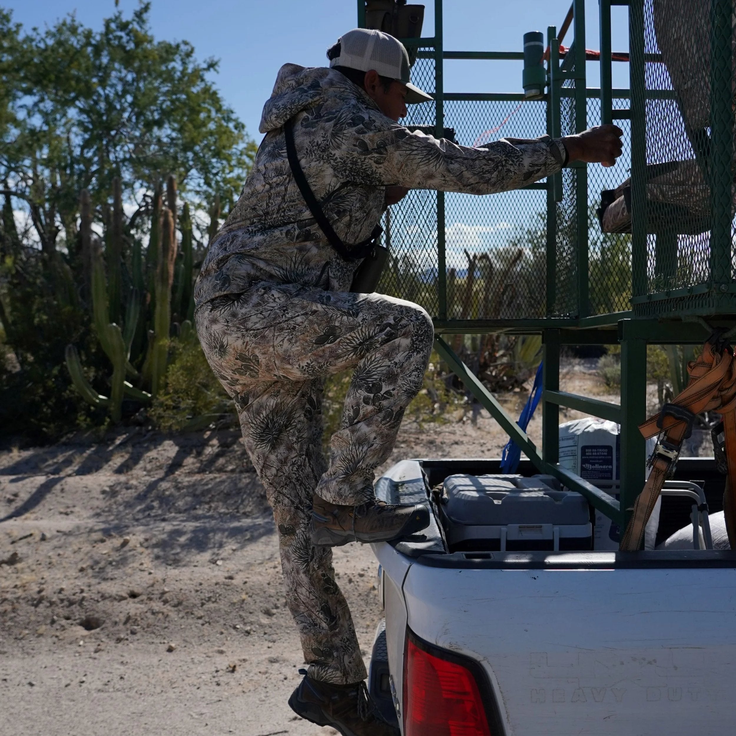 Man in desert camo hunting suit climbing into truck bed, cactus backdrop, outdoor gear ready