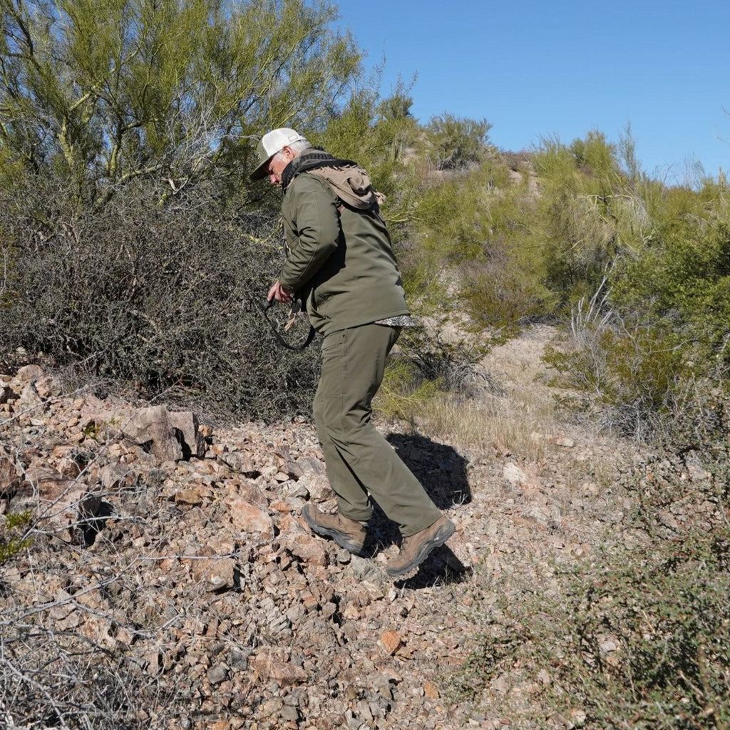 Man in olive green hunting jacket and pants trekking rocky desert terrain with backpack and cap