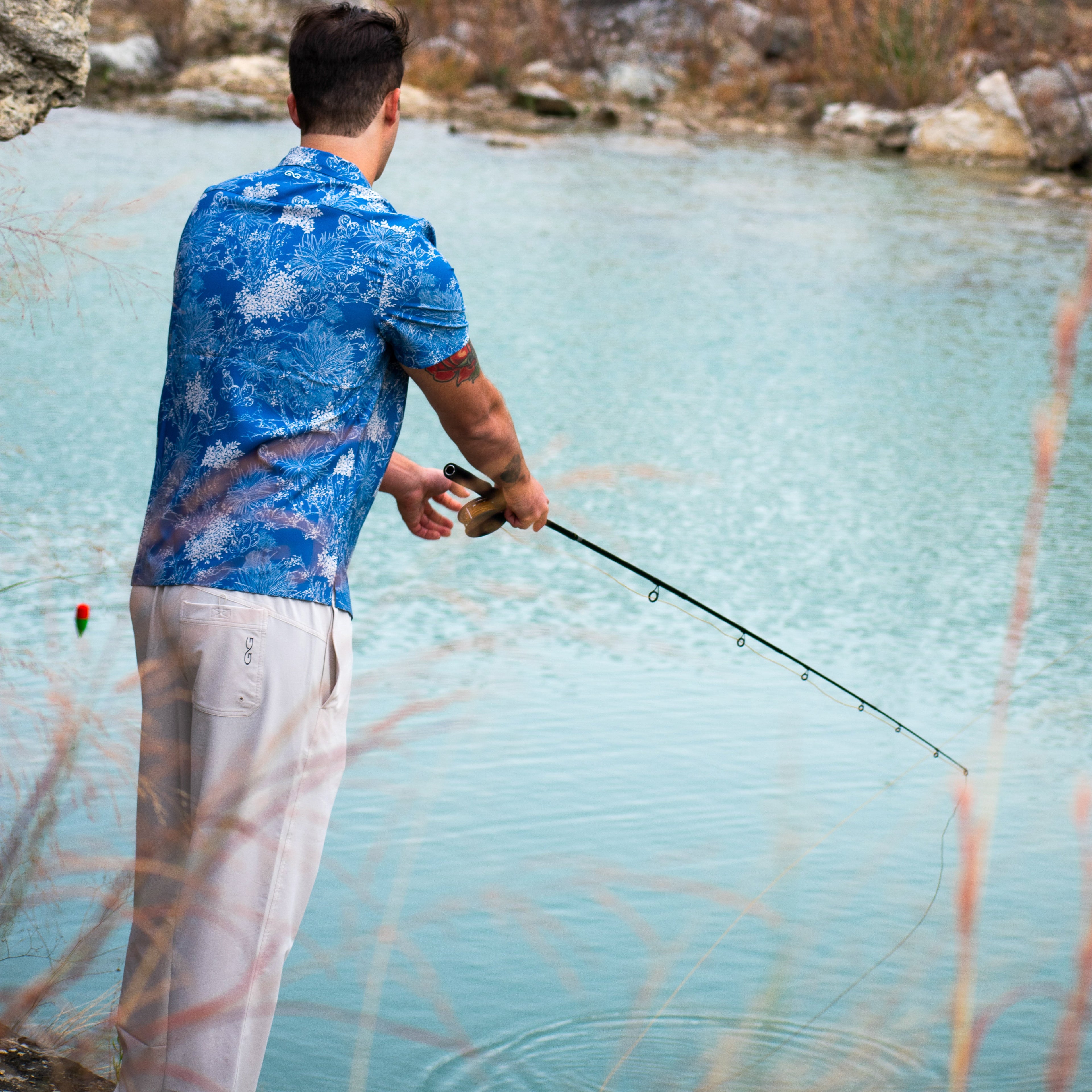 Man fishing in blue floral shirt and beige pants by river, holding rod with float