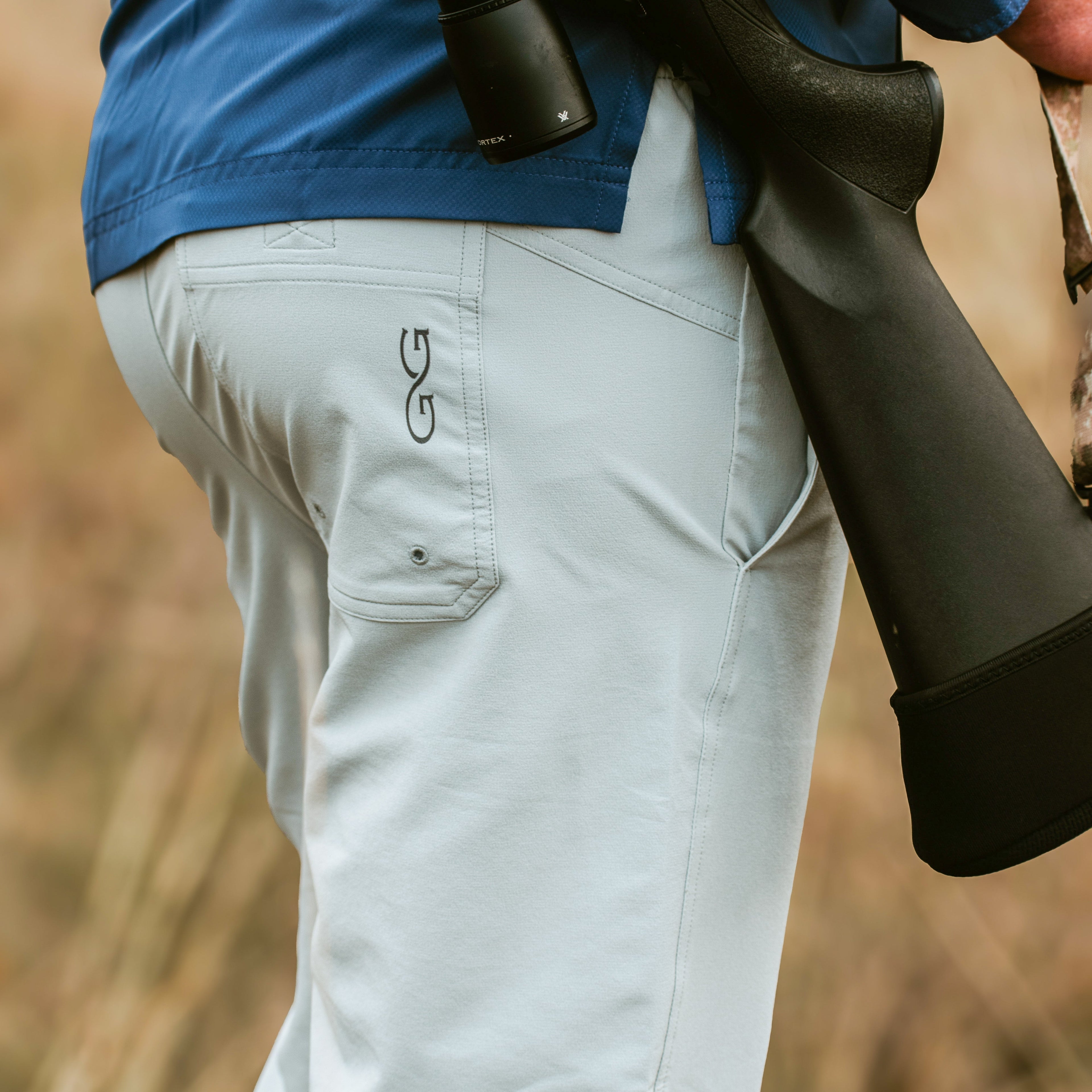 Light gray hunting pants with logo, worn with blue shirt and shotgun, ideal for outdoor adventures