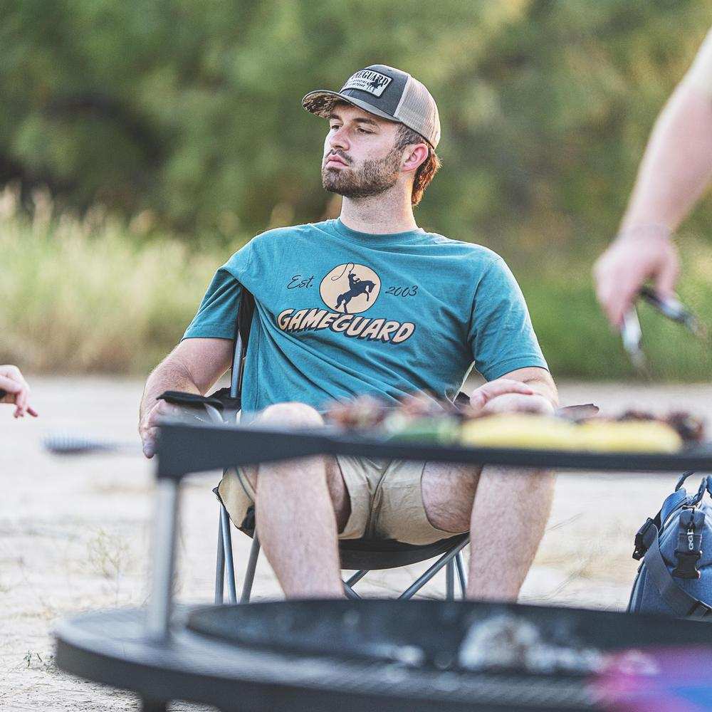 Man wearing teal GameGuard t-shirt and camo trucker hat outdoors by campfire, hunting apparel