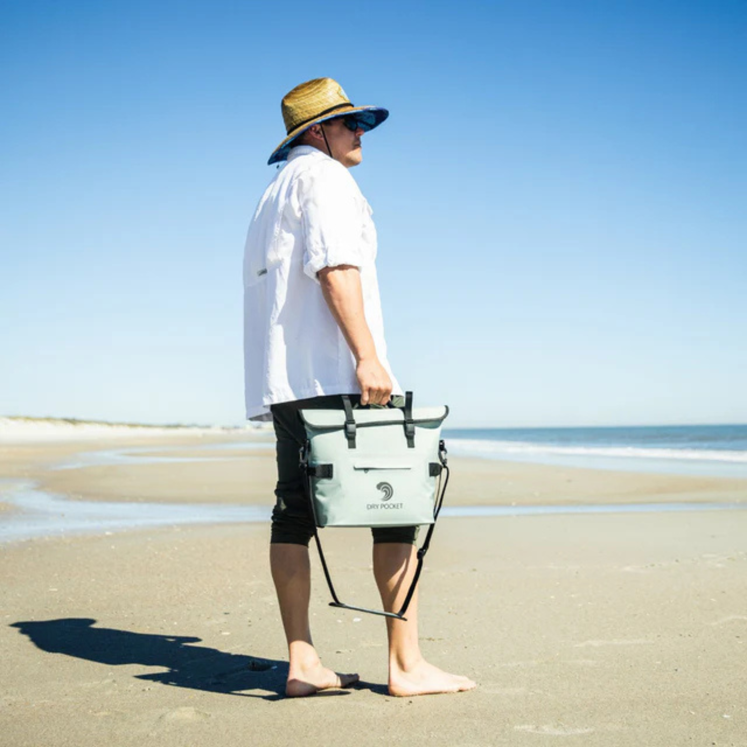 Man on beach holding light green Dry Pocket waterproof gear bag for fishing or hunting adventures