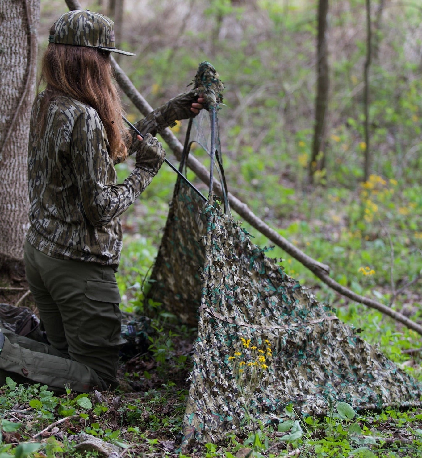 Woman setting up woodland camo hunting blind in forest, wearing matching camo gear and gloves