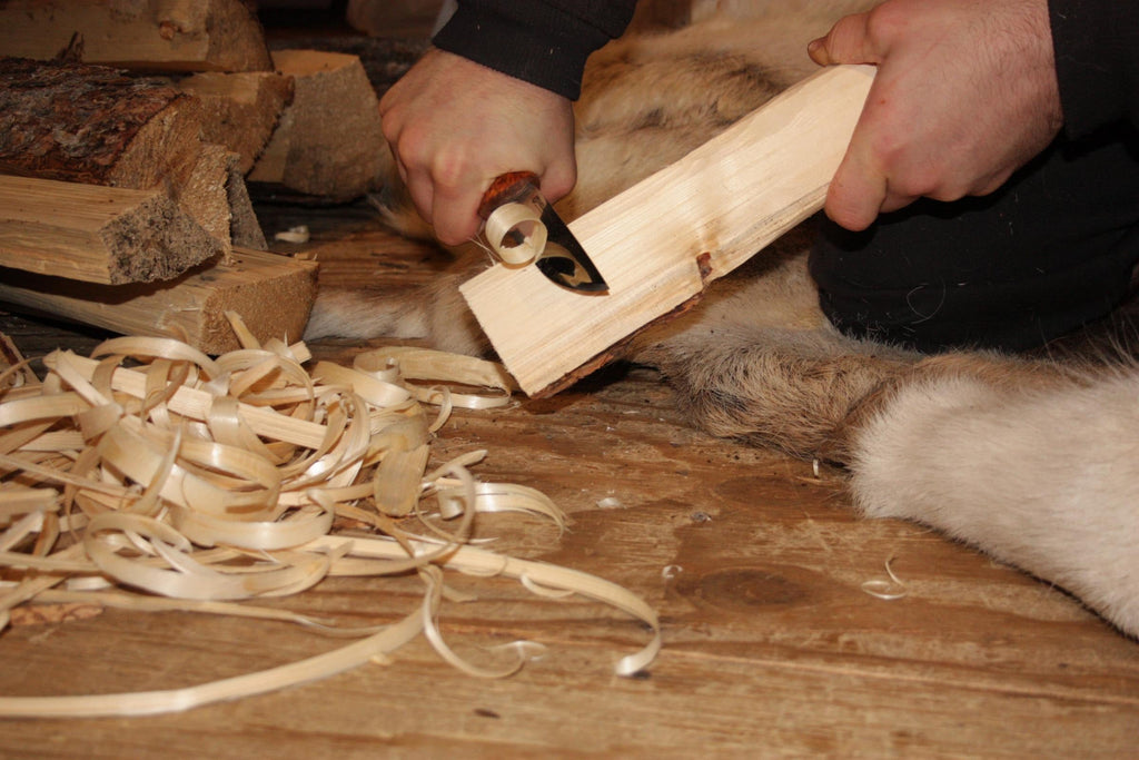 Person carving wood with knife on fur rug, outdoor survival gear